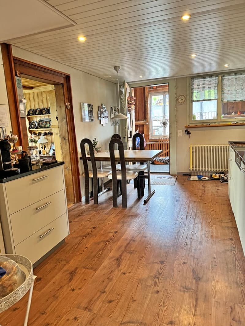 kitchen with dining area, wooden floor, dining table with chairs, kitchen counter with appliances, shelves with decorative items, radiator, windows with curtains