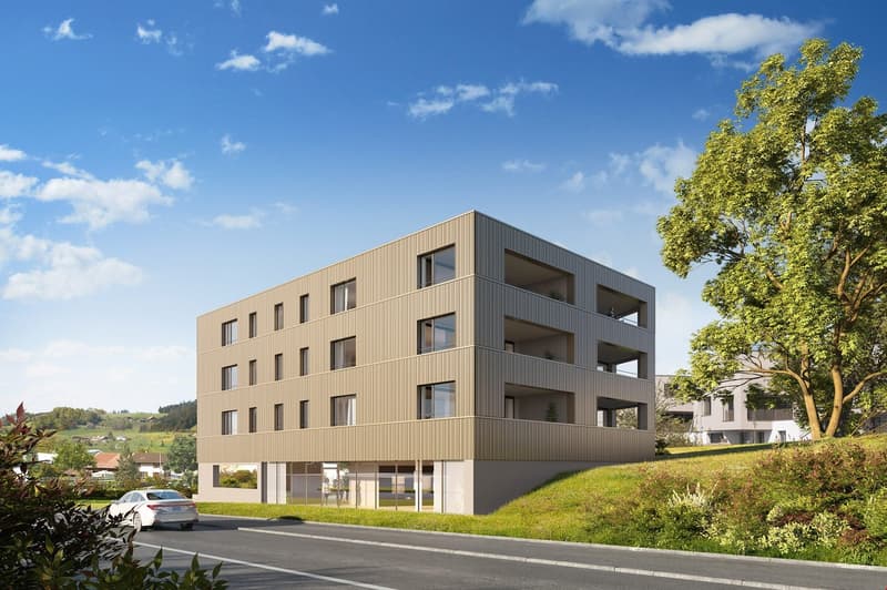 3-story apartment building with a modern, minimalist design featuring a tan/beige exterior cladding and large windows. The building has balconies on each floor and is situated on a grassy slope with trees in the background.