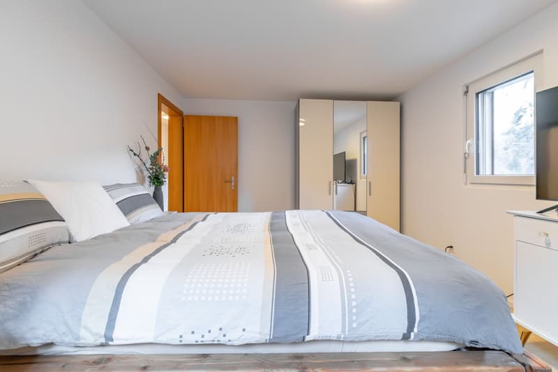 bedroom with wooden floor, white walls, large bed, wooden door, wardrobe with mirror, tv, flower vase, night stand