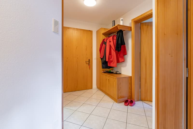 entryway, wooden door, tiled floor, coat rack with clothes, storage box, red slippers