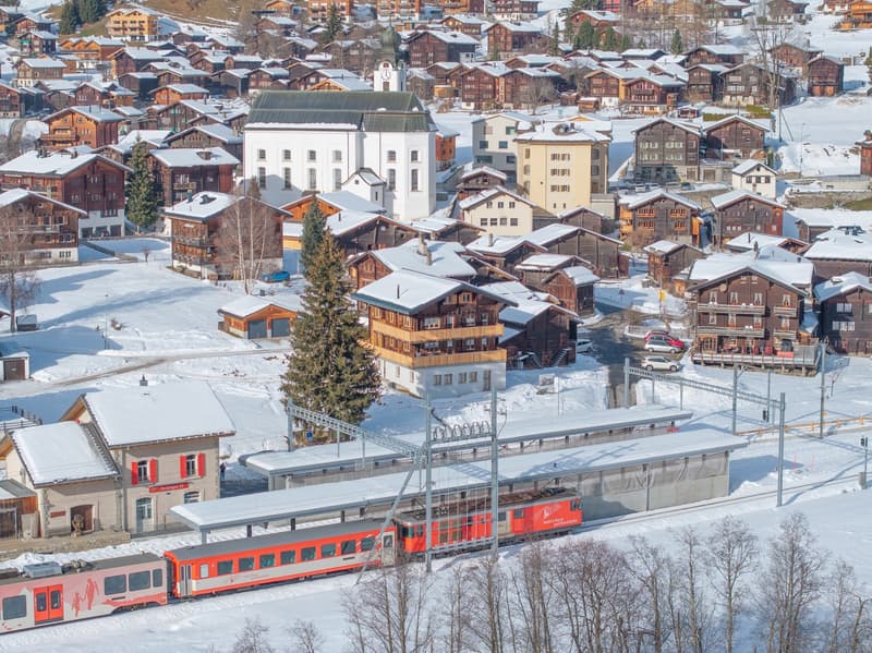 Aerial view of the town of Grindelwald, Switzerland covered in snow. Multiple wooden chalets, a church, and a train station. The train is stopped and partially covered in snow. Snow-covered mountains in the distance.