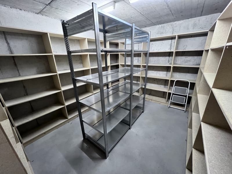 The image shows a storage room with multiple metal shelving units. The shelves are empty and the room has a concrete floor and walls. There are also a couple of small black stools visible in the corner.