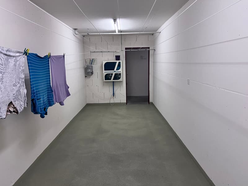 Narrow hallway with concrete floor, white walls, and a washing machine mounted on the wall. Clothes are hanging on a clothesline.