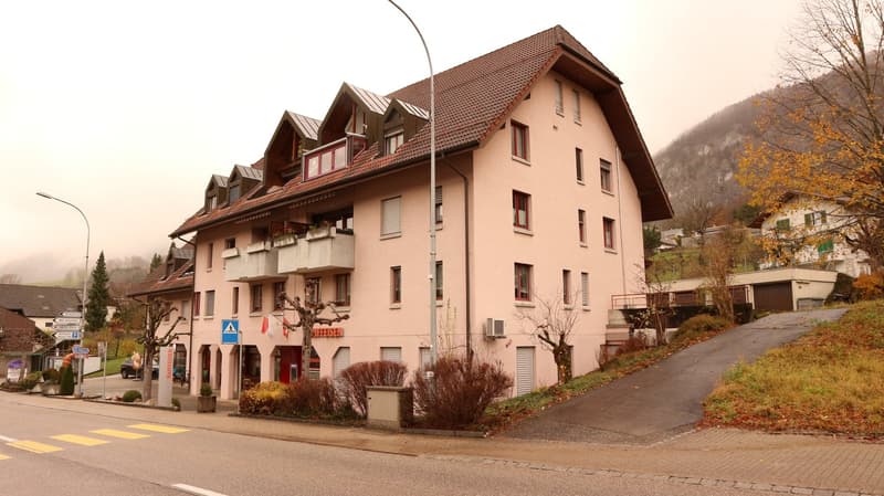 Multi-story apartment building with red tiled roof, pink exterior walls, and balconies. The building is situated in a mountainous area with trees and a road in the foreground.
