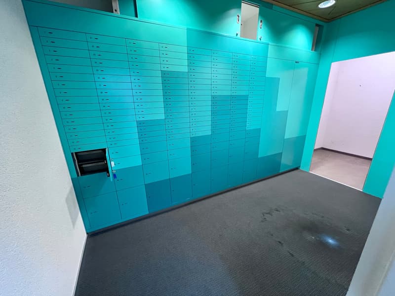 The image shows a room with numerous teal-colored lockers or storage units lining the walls. The floor is covered with a dark gray, textured material. There is a doorway visible on the right side of the image.