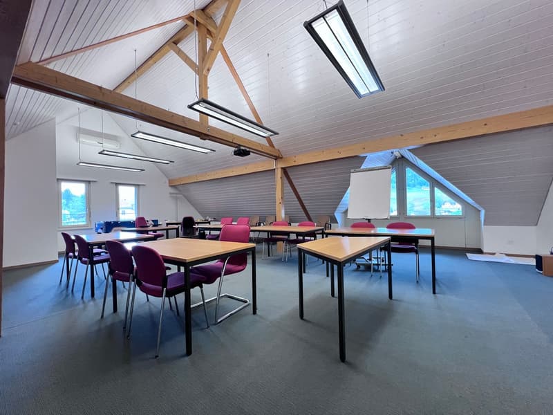 The image shows a large classroom or study area with wooden beams and a high ceiling. The room has multiple tables and chairs arranged in rows, suggesting it is used for educational or meeting purposes. The walls are white and the floor is a dark gray col