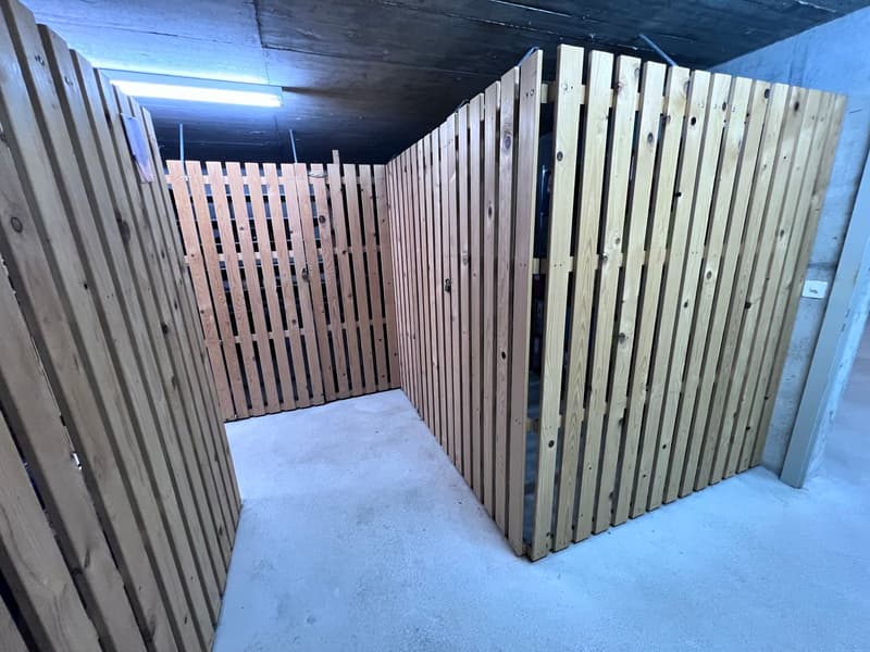 The image shows a storage unit or locker area with wooden slat walls and a concrete floor. The space appears to be divided into individual storage compartments or lockers, with wooden slats forming the walls and doors. The overall structure has a utilitar
