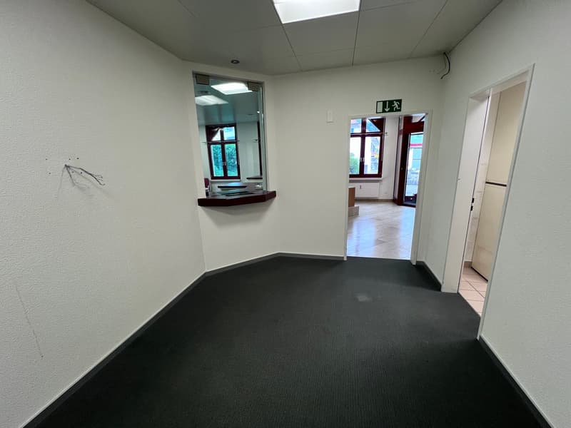 The image shows a small office space with white walls, a carpeted floor, and a window. There is a small shelf or ledge on the wall and a doorway leading to another room.