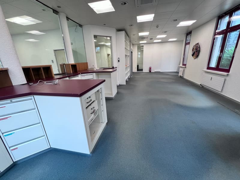 The image shows a large, open office space with a reception area, storage cabinets, and a long hallway leading to other rooms. The space has a modern, professional design with gray carpeting, white walls, and recessed lighting. There are no visible featur