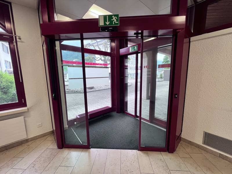 The image shows the entrance of a building with automatic sliding glass doors. The doors are framed in a burgundy-colored metal structure, and there is an emergency exit sign above the doors. The floor inside the entrance is covered with gray carpeting.