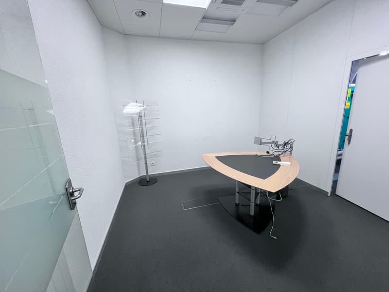Small office space with white walls, black carpeted floor, a curved wooden desk, and a standing lamp. The room also has a glass door and appears to be equipped with air conditioning.