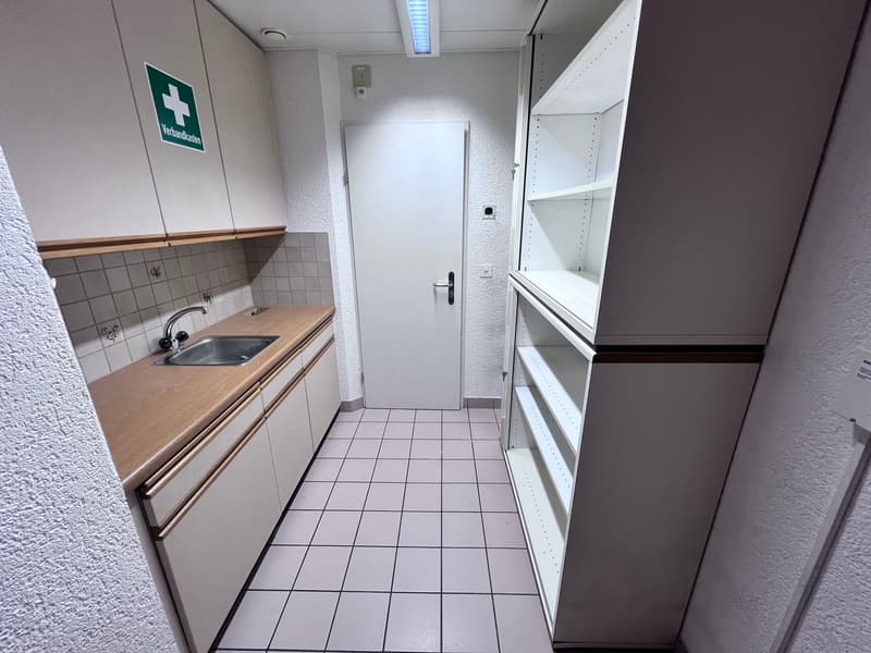 Small kitchenette with a sink, cabinets, and a refrigerator. Tiled floor and walls. Emergency first aid sign on the wall.