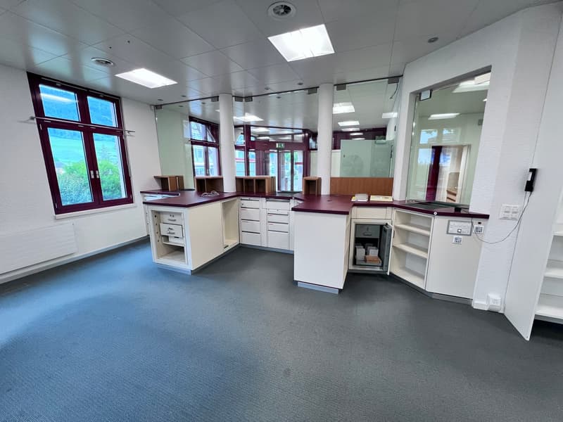 The image shows a modern, well-lit office space with a reception area. The room has white walls, a gray carpet floor, and large windows that provide natural light. The space is equipped with various office furniture and equipment, including desks, drawers