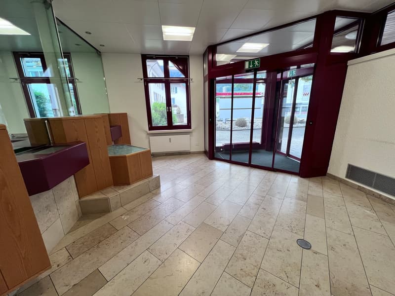 The image shows a commercial office space with a tiled floor, wooden cabinets, and large windows. The space appears to be an entrance or lobby area with a clean and modern design.