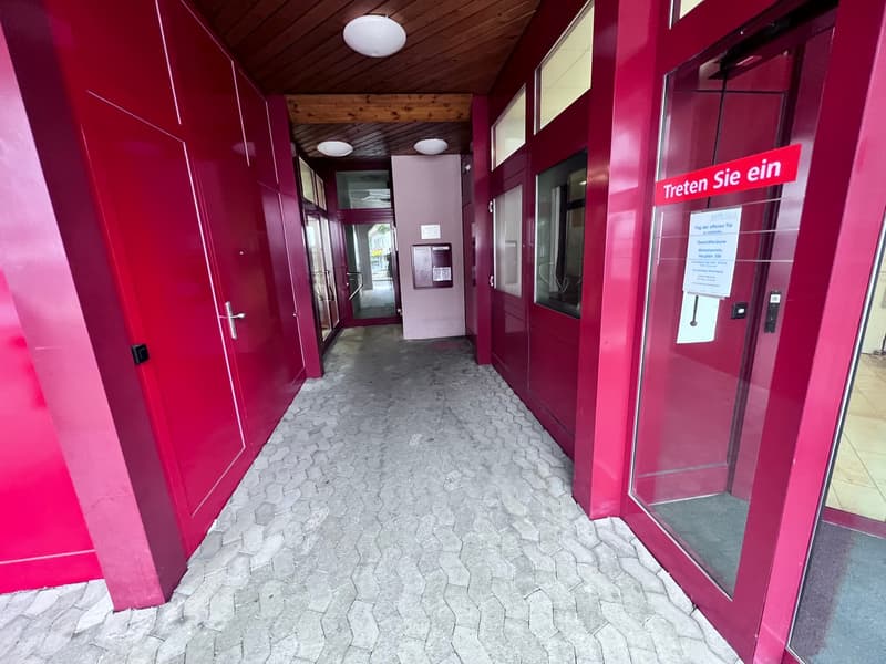 The image shows a hallway with bright pink walls and a wooden ceiling. The floor is made of gray hexagonal tiles. There are glass doors leading to the outside and a sign in German that says 'Enter here'.