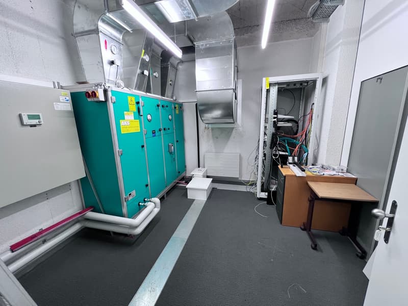 The image shows a utility room or service area with various equipment and machinery. There is a large green cabinet or unit, likely an air conditioning or ventilation system. The room also contains electrical panels, wiring, and other equipment. The floor
