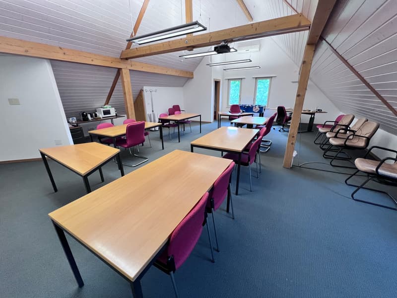 The image shows a classroom-like setting with several wooden tables and purple chairs arranged in rows. The room has a high ceiling with exposed wooden beams and a gray paneled ceiling. There are windows on one side of the room, and various equipment and