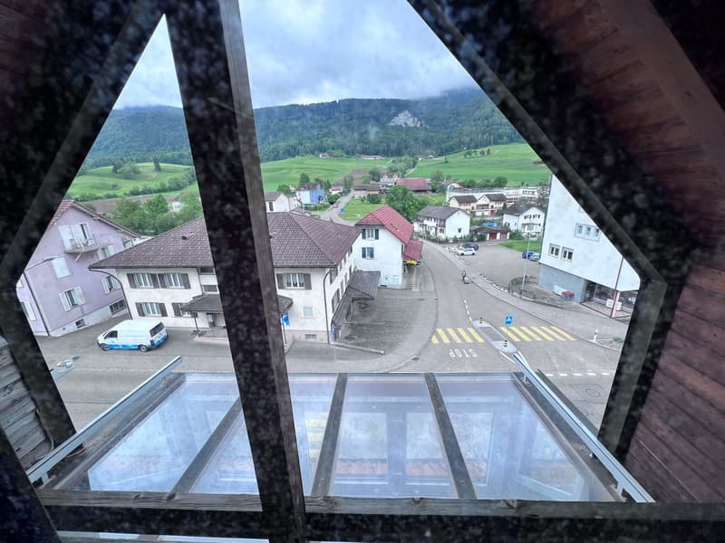 View of a small town or village surrounded by mountains and green hills, with residential buildings, roads, and a few vehicles visible. The image is captured through a glass window or structure, providing a panoramic view of the scenic landscape.
