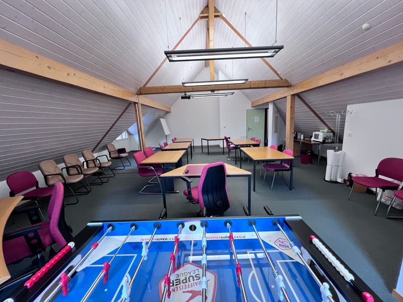 Large open classroom with high ceilings, wooden beams, and a foosball table in the foreground. The room is equipped with multiple desks and chairs, as well as a kitchenette area.