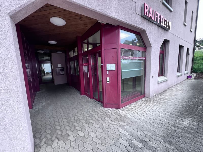 The image shows the exterior of a building with a pink/purple color scheme. The building has a glass entryway and the name 'Raiffeisen' is displayed prominently on the facade. The entrance is surrounded by a paved walkway with a hexagonal pattern. The ove