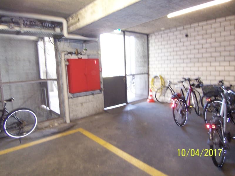 The image shows a garage or storage space with a red storage cabinet, several bicycles parked, and a concrete floor with a yellow line marking the space.