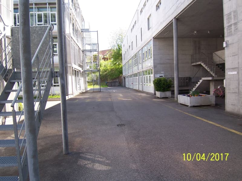 The image shows a commercial or institutional building with a concrete exterior and multiple floors. There are stairs and railings leading up to the building, and the surrounding area appears to be a paved walkway or courtyard. The building has a modern,