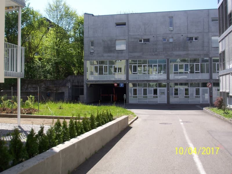 A multi-story concrete building with large windows, surrounded by trees and greenery. There is a parking area in front of the building.
