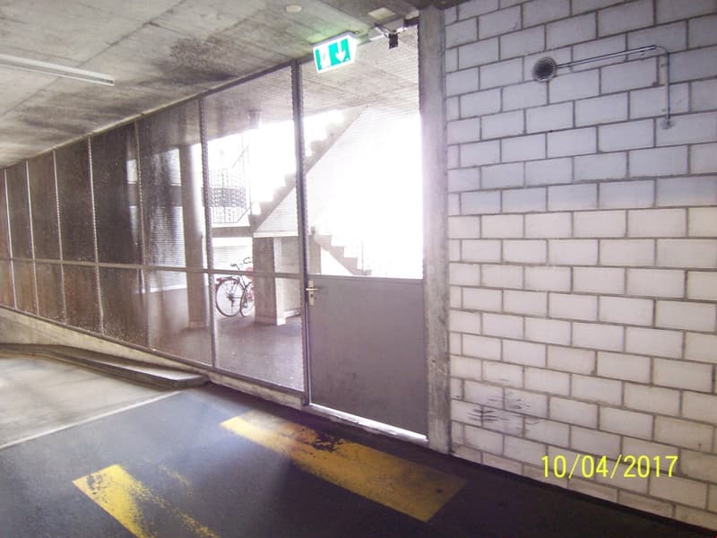 The image shows a hallway or corridor with white brick walls, a glass partition, and a staircase visible through the glass. There is an exit sign and a bicycle visible in the background.