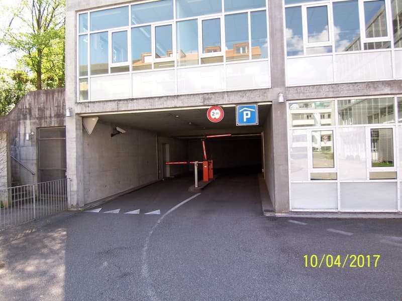 The image shows the entrance to an underground parking garage. The entrance is marked with a parking sign and has a 2m height restriction. The garage appears to be located in a commercial or office building, with a glass facade visible above the entrance.
