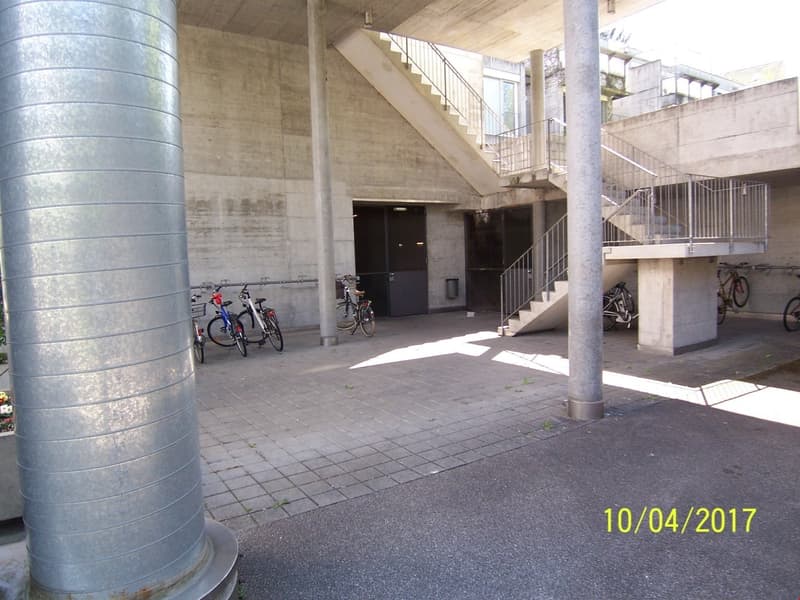 The image shows the entrance of a building with a concrete structure and stairs leading up to the entrance. There are several bicycles parked in the area, indicating a parking space for bicycles. The building appears to be a multi-story structure with a m