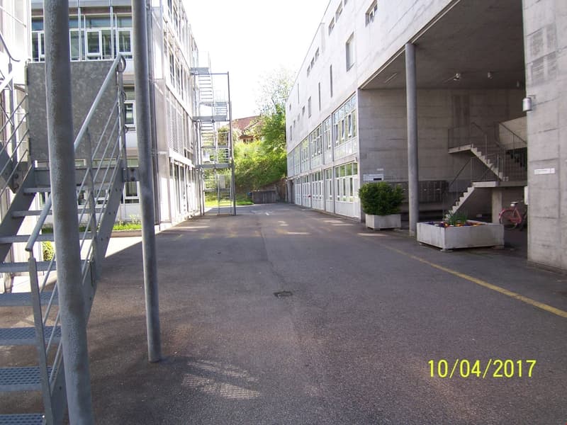 The image shows a commercial or institutional building with multiple floors and a concrete exterior. There are stairs and railings leading to the entrance, and the building appears to be surrounded by a paved area or courtyard. The overall scene suggests