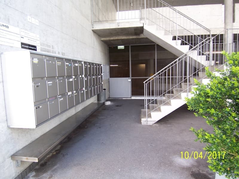 The image shows the entrance to a building, with a set of metal lockers or mailboxes on the left side and a staircase leading up to the second floor on the right. The entrance appears to be made of concrete and has a metal railing. There are some plants v