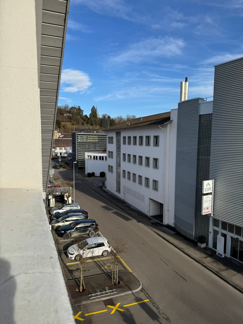 The image shows a multi-story commercial or office building with a parking lot in the foreground. The building has a modern, industrial design with gray and white facades. There are several cars parked in the lot, and the sky is clear with some clouds vis