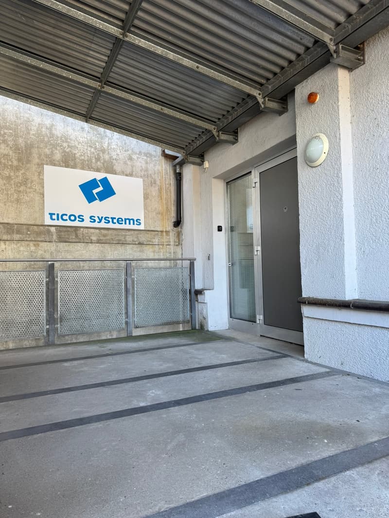 The image shows the exterior of a commercial building belonging to Ticos Systems. The building has a metal roof structure and a gray exterior wall. There is a sign with the Ticos Systems logo on the front of the building, and a glass door entrance. The gr