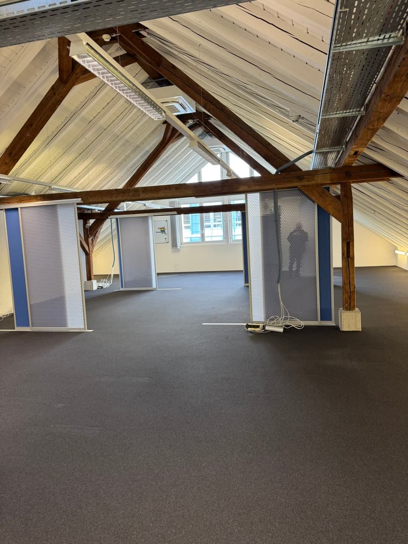 The image shows a large open space with a high, exposed wooden ceiling and beams. The walls are painted in a light color, and the floor is covered with gray carpeting. There are several windows along the walls, providing natural light. The space appears t