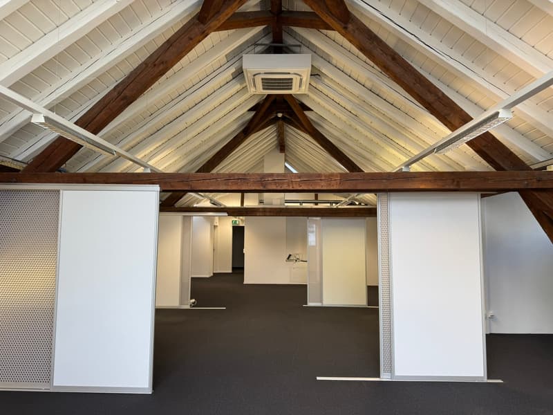 Large open office space with high vaulted ceilings, exposed wooden beams, and a fireplace. The space has white walls, dark carpeted floors, and several partitioned areas.