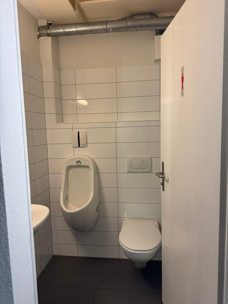 The image shows a bathroom with white tiled walls, a urinal, a toilet, and a ventilation duct running along the ceiling.