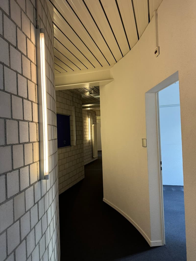 The image shows a hallway with brick walls, a vaulted ceiling with wooden beams, and a doorway leading to another room. The hallway has a tiled floor and is well-lit with a light fixture on the ceiling.