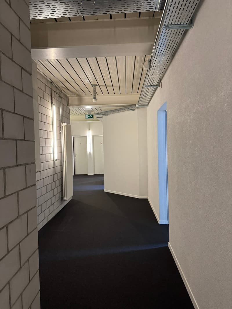 The image shows a hallway with white tiled walls, a suspended ceiling with horizontal slats, and a dark floor. The hallway has two doors visible, indicating it is a common area or corridor in a building.