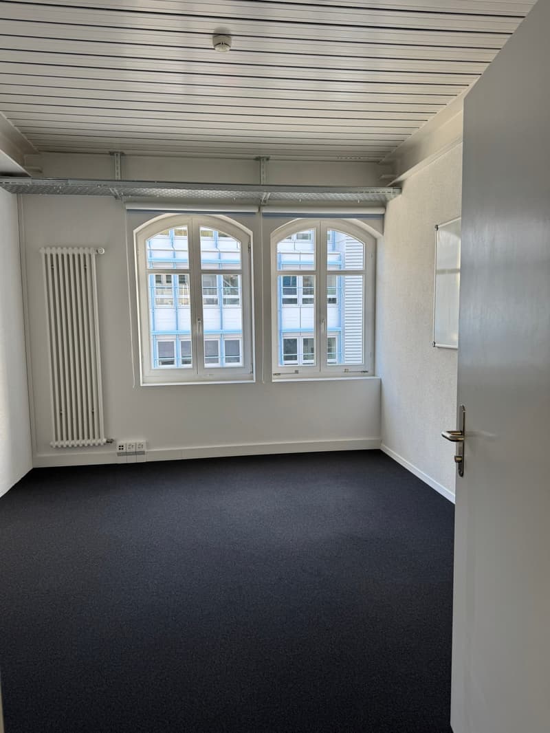 The image shows an empty room with white walls, a gray ceiling with horizontal panels, and large arched windows that let in natural light. The floor is covered with a dark gray carpet. The room appears to be a simple, minimalist space, likely intended for
