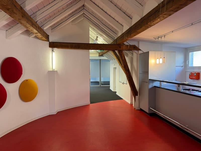 Open-concept loft-style space with exposed wooden beams, high ceilings, and a mix of modern and colorful decor elements. The space includes a kitchen area with stainless steel appliances, and a living/work area with red and yellow circular wall decoration