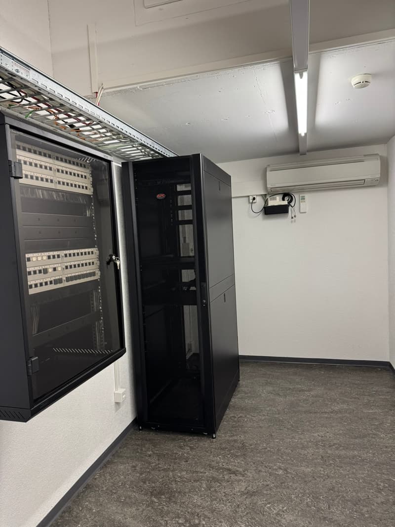The image shows a server room with a large server rack and a separate server cabinet. There is also an air conditioning unit visible on the wall.