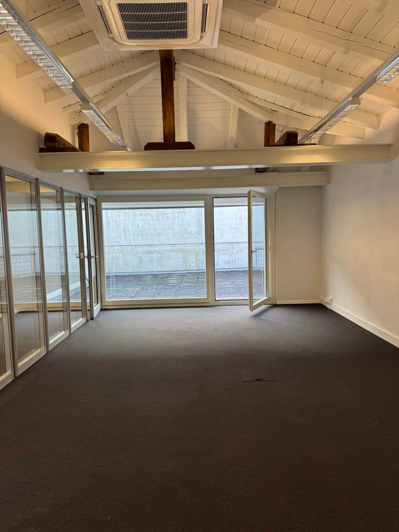 Large open space with high ceilings, wooden beams, and sliding glass doors leading to a balcony or terrace. The room has a carpeted floor and an air conditioning unit installed in the ceiling.