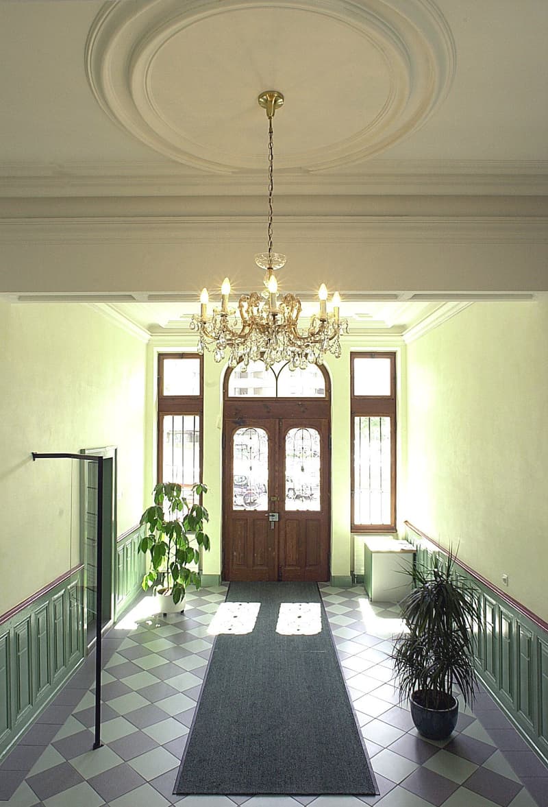Spacious entrance hall with a grand chandelier, ornate ceiling, wooden double doors, checkered tile floor, and potted plants.