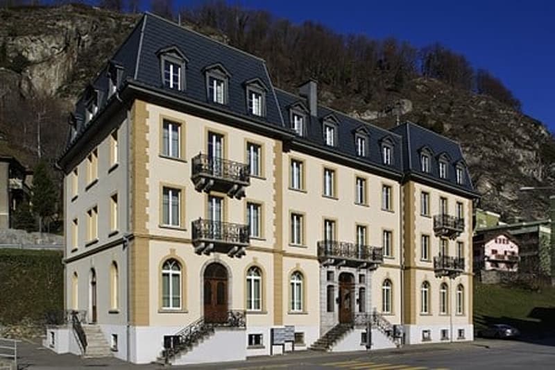 A multi-story building with a yellow facade, black roof, and balconies. The building appears to be located in a mountainous area with trees and a blue sky in the background.