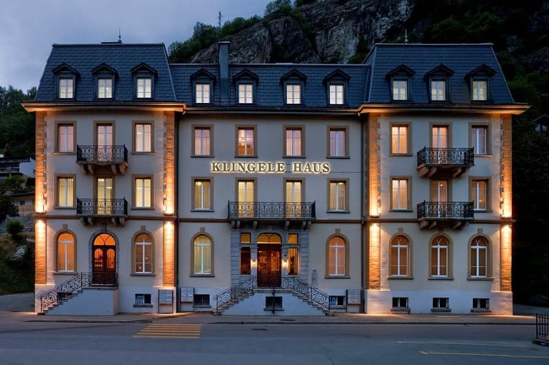 A multi-story hotel building with a traditional European architectural style, featuring stone walls, balconies, and large windows. The building is illuminated at night, creating a warm and inviting atmosphere. The name 'Klingele Haus' is prominently displ