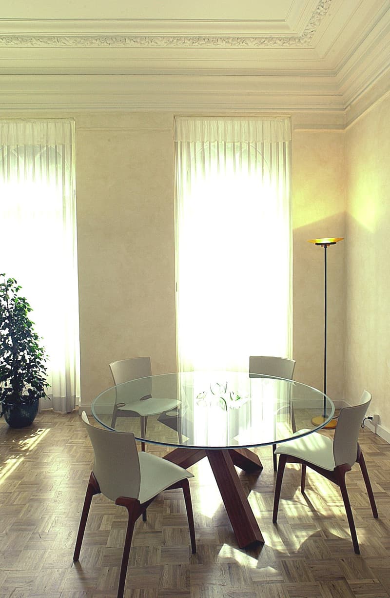 The image shows a dining room with a round glass table and four chairs. The room has high ceilings with decorative molding, and large windows with white curtains that allow natural light to fill the space. There is a potted plant in the corner, adding a t