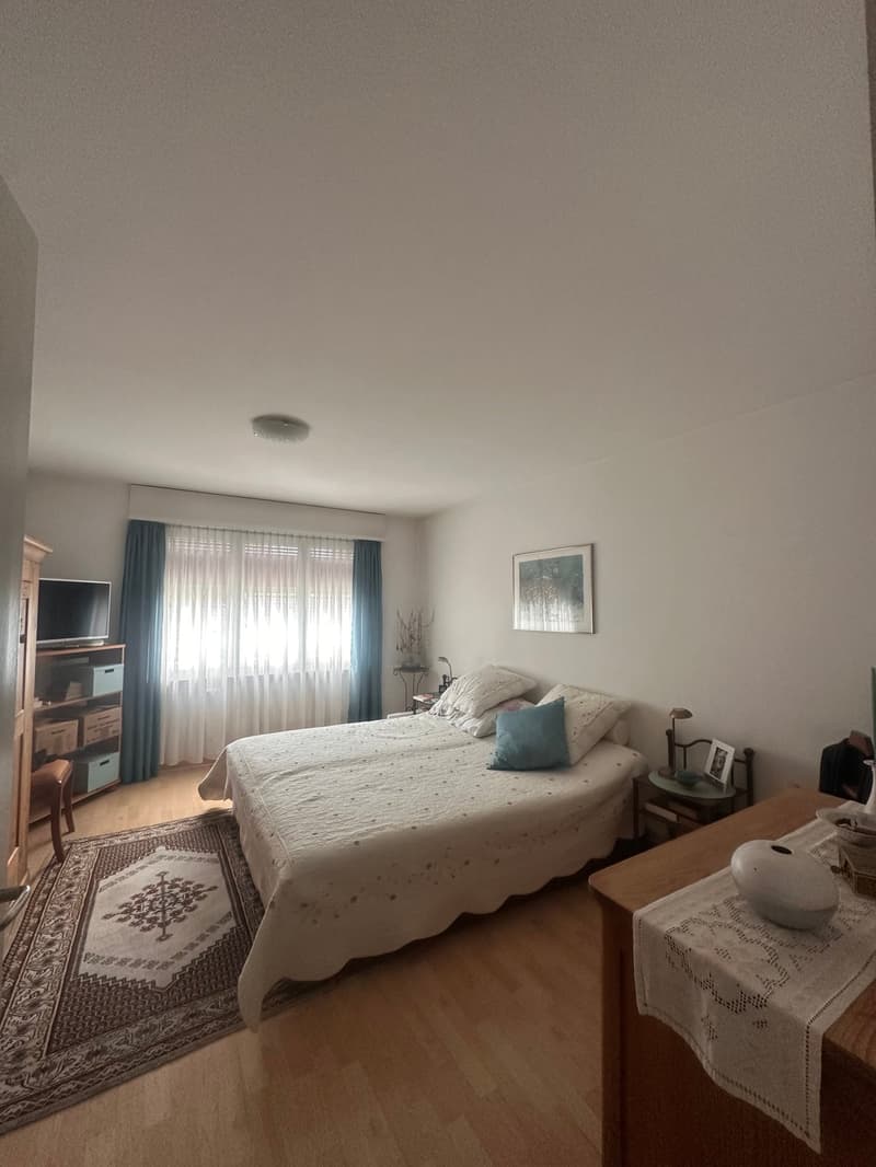 Bedroom with a double bed, wooden floor, white walls, a rug, a cabinet with a TV, a window with blue curtains, a picture frame, a nightstand with a lamp, and a table with a white cloth and a bowl.