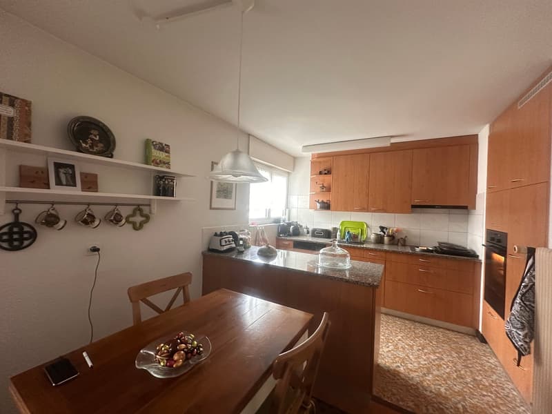 kitchen with dining area, wooden table, chairs, wooden cabinets, granite countertop, kitchen appliances, white walls, ceiling lamp, shelves with decorative items