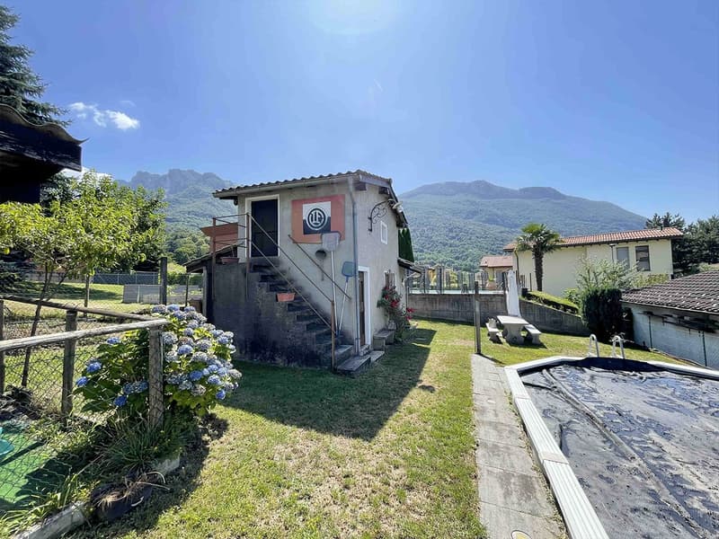 Detached house with staircase, wooden door, logo on the wall, outdoor area with a covered terrace, table, and bench, surrounded by a fence, lush vegetation, mountains in the background